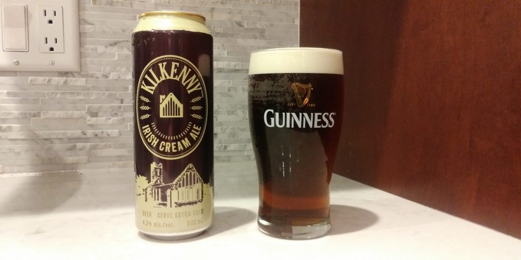 An empty can of Kilkenny nitro brew cream ale sits next to a full glass marked with the Guinness nitro brew stout logo on white counter with a white tile and dark wood background.